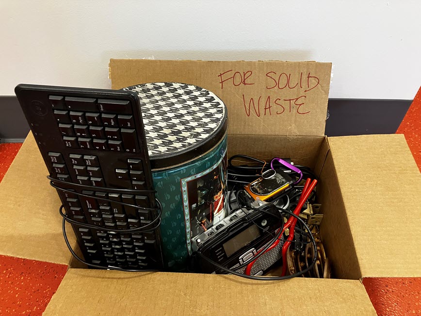 box of small electronic items (keyboard, mouse, radio) and random metal items set out next to recycling cart labeled 'for solid waste'