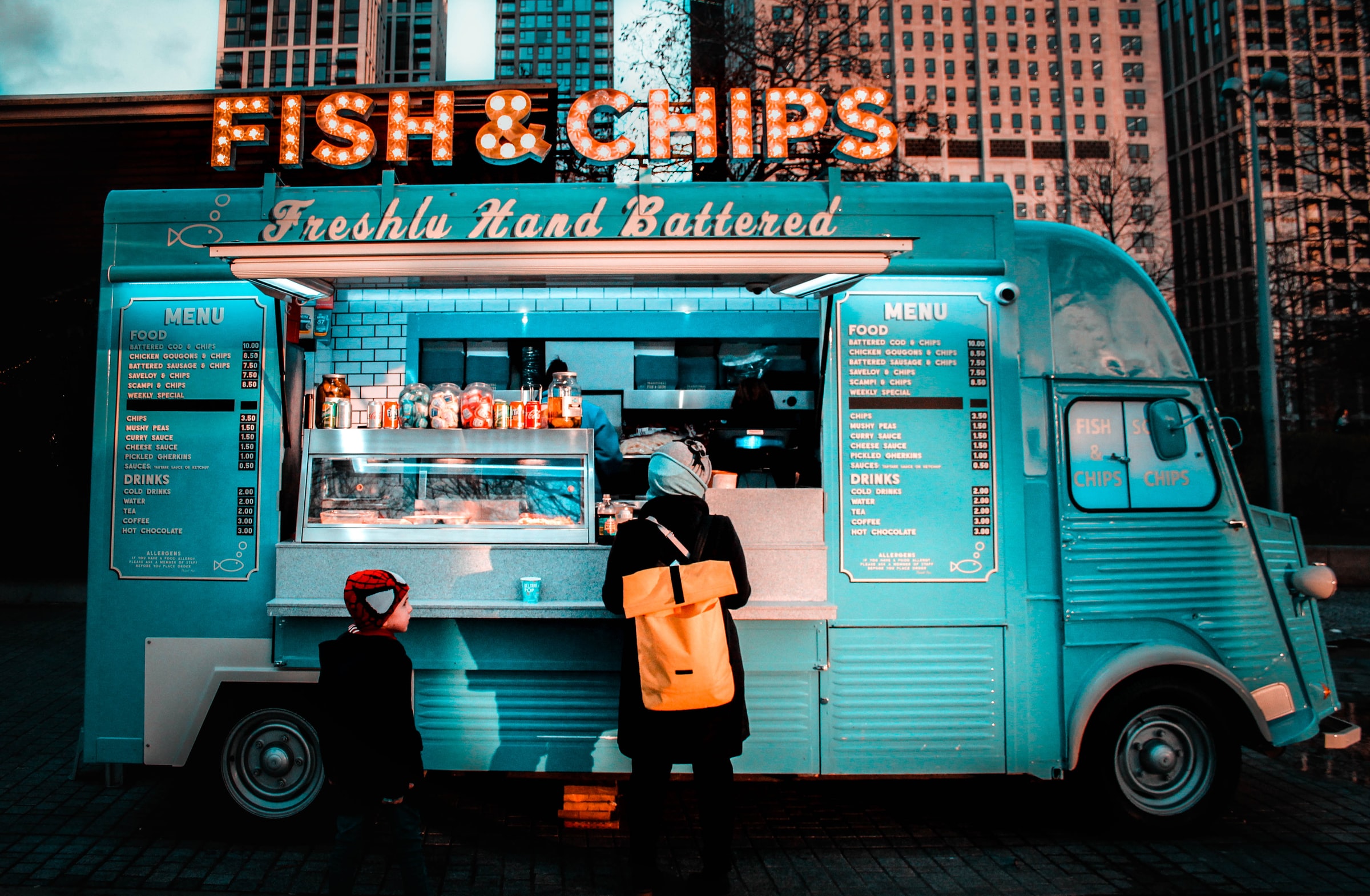 Blue food truck with orange neon Fish & Chips sign on top and one adult and one child customer facing the truck.